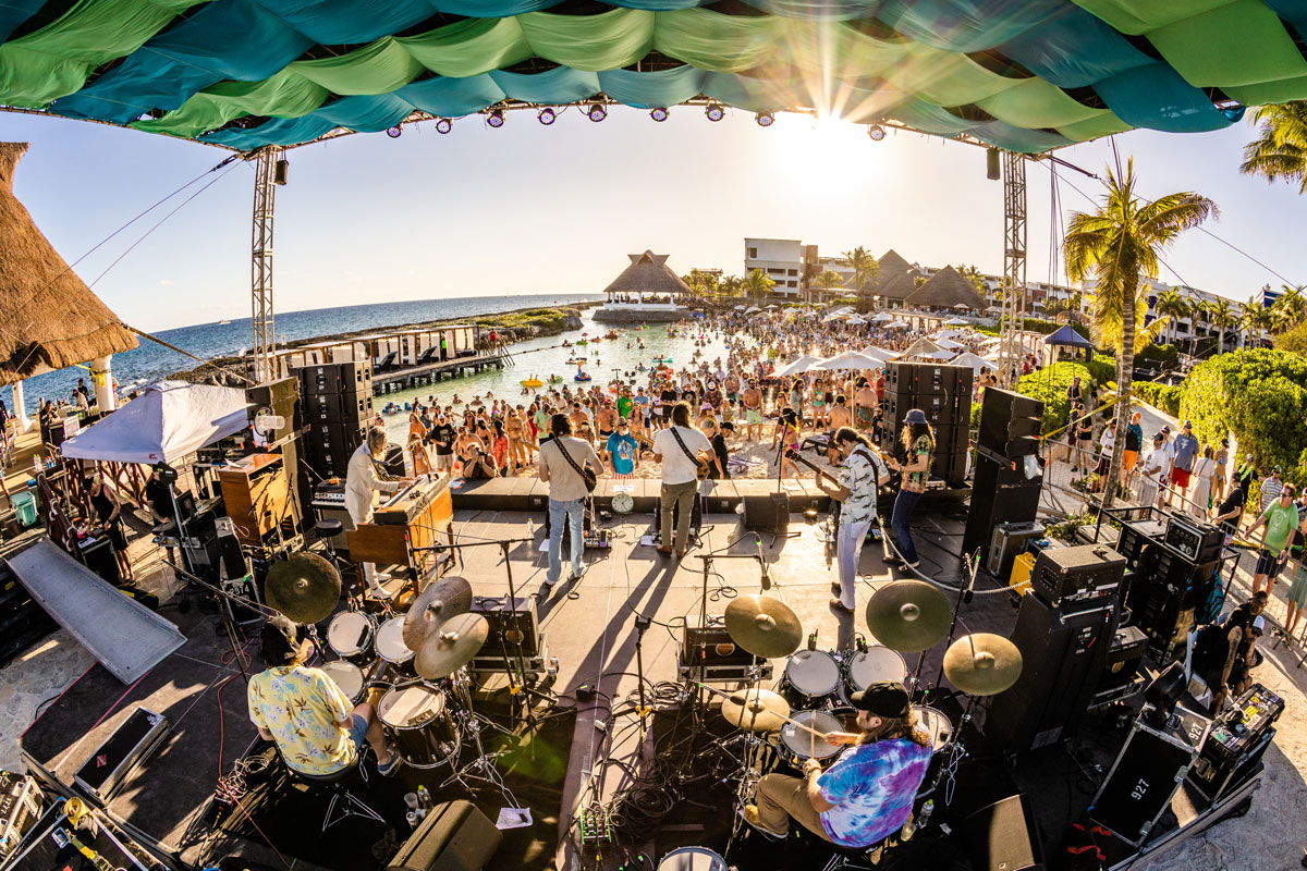 Photo from behind a band performing on the beach stage, crowd watching while floating in a lagoon.